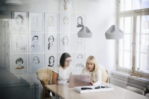 Viewed from behind an office window which shows slight reflections, two women are sitting at a table discussing something that is on a laptop in front of them. The office wall behind them is covered in stylised, illustrated portraits.