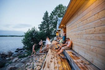 On the right a small group of people are chilling outside of the Lonna sauna on Lonna island, a few trees at the back of the building, and the shoreline stretching into the distance to the left of the photo.