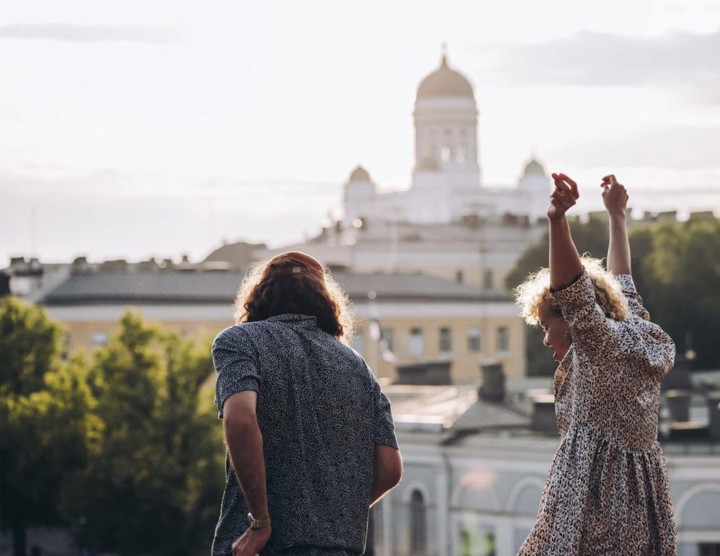 With their backs facing the camera and standing on a hill in front of Uspenski Cathedral, the man on the left faces forward while the woman on the right reaches her arms into the air whilst smiling. In the background, rooftops and trees can be seen, Helsinki Cathedral sitting on the horizon.