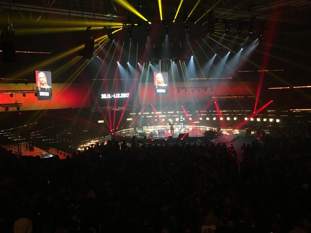 A large crowd are seated in darkness around the main stage at Slush, a speaker addresses the audience while dozens of different coloured spotlights light up the scene.