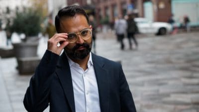 With Stockmann standing behind in a blurred background, Sameer Narula, participant in 90 Day Finn, stands looking towards the camera with one hand raised and holding his glasses.