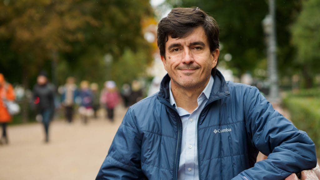 Jaime Alvarez - 90 Day Finn, sitting on a brown bench in the Esplanadi park, in the background green trees and people can be seen in a blurred way, Jaime is smiling at the camera and wearing a light blue shirt and a dark blue jacket