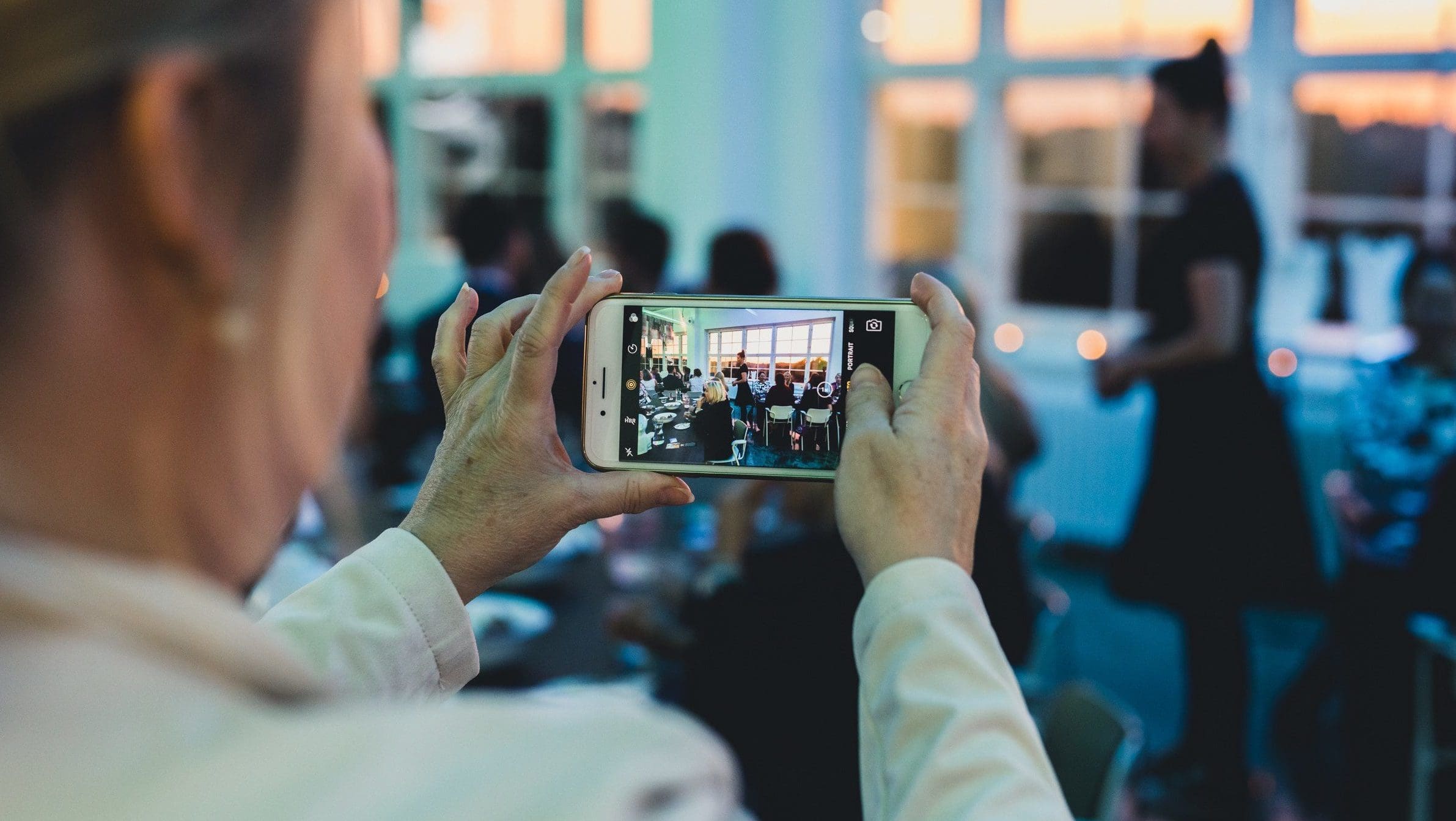 A ladi is taking a landscape picture of a group of people. The people in the pictures are dressed as in a formal business event, they either sit at the table or stand up and discuss among themselves. The lady who is taking the picture is photographed from her back. She seems to be dressed with a white shirt and have a lanyard around the neck.