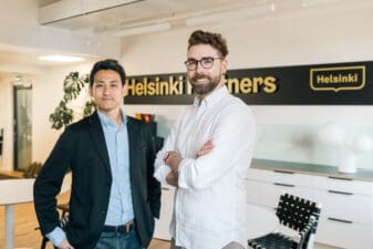The two partners from NordicNinja (men) are standing and looking at the camera. Behind them there is the Helsinki Partners logo and the Helsinki Partners office background. One of the man is from Japan and is wearing a black suit and a blue shirt, the other man is from Finland and he wears a white shirt.