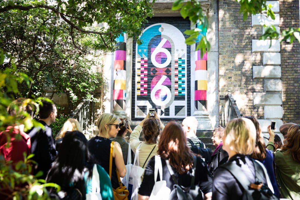A design event by Media 10. People looking at a colourful artwork under leafy trees.