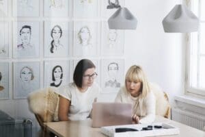 Viewed from behind an office window which shows slight reflections, two women are sitting at a table discussing something that is on a laptop in front of them. The office wall behind them is covered in stylised, illustrated portraits.